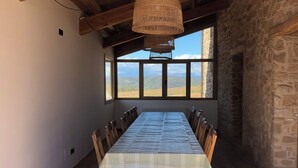 Un comedor con una larga mesa de madera, paredes de piedra y grandes ventanales con vistas al paisaje.