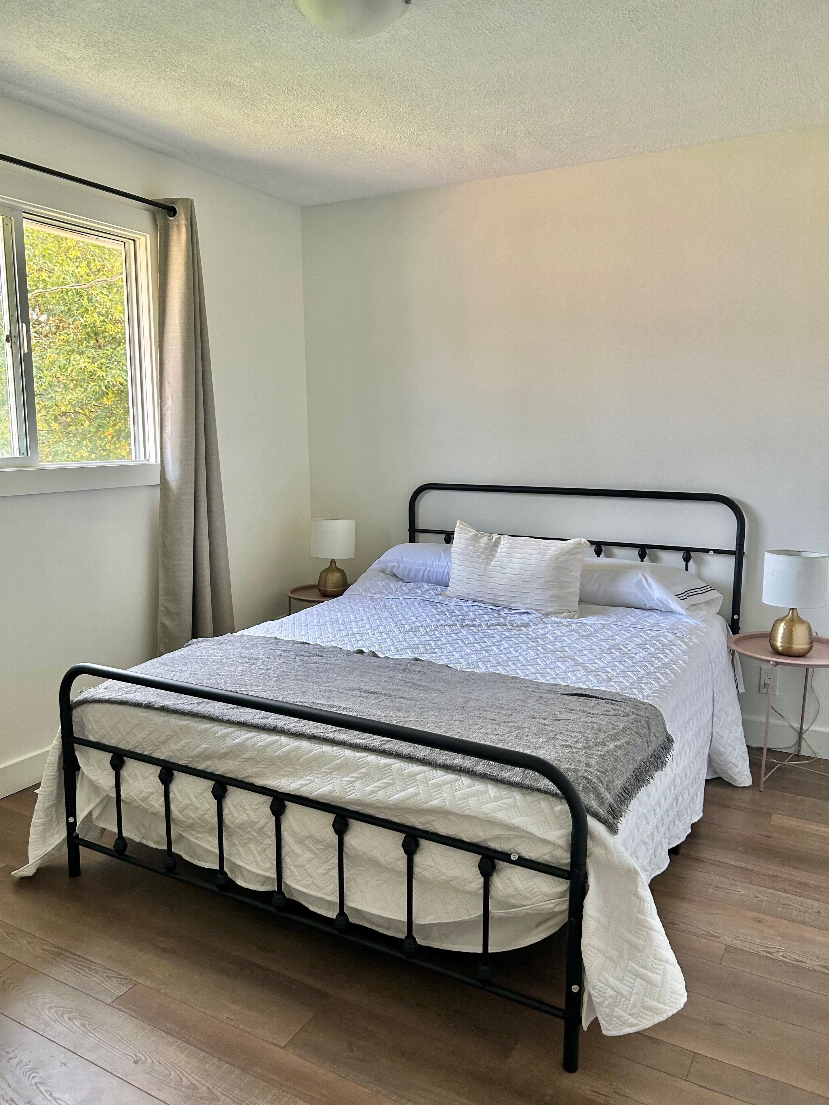 Una cama bien hecha con estructura metálica, una mesita de noche con lámpara y una ventana con cortinas.