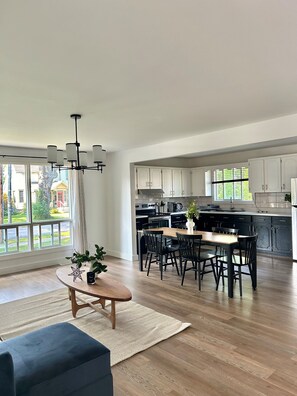 A spacious kitchen with modern appliances, a dining area with a wooden table and chairs, and large windows offering a view of the outside.