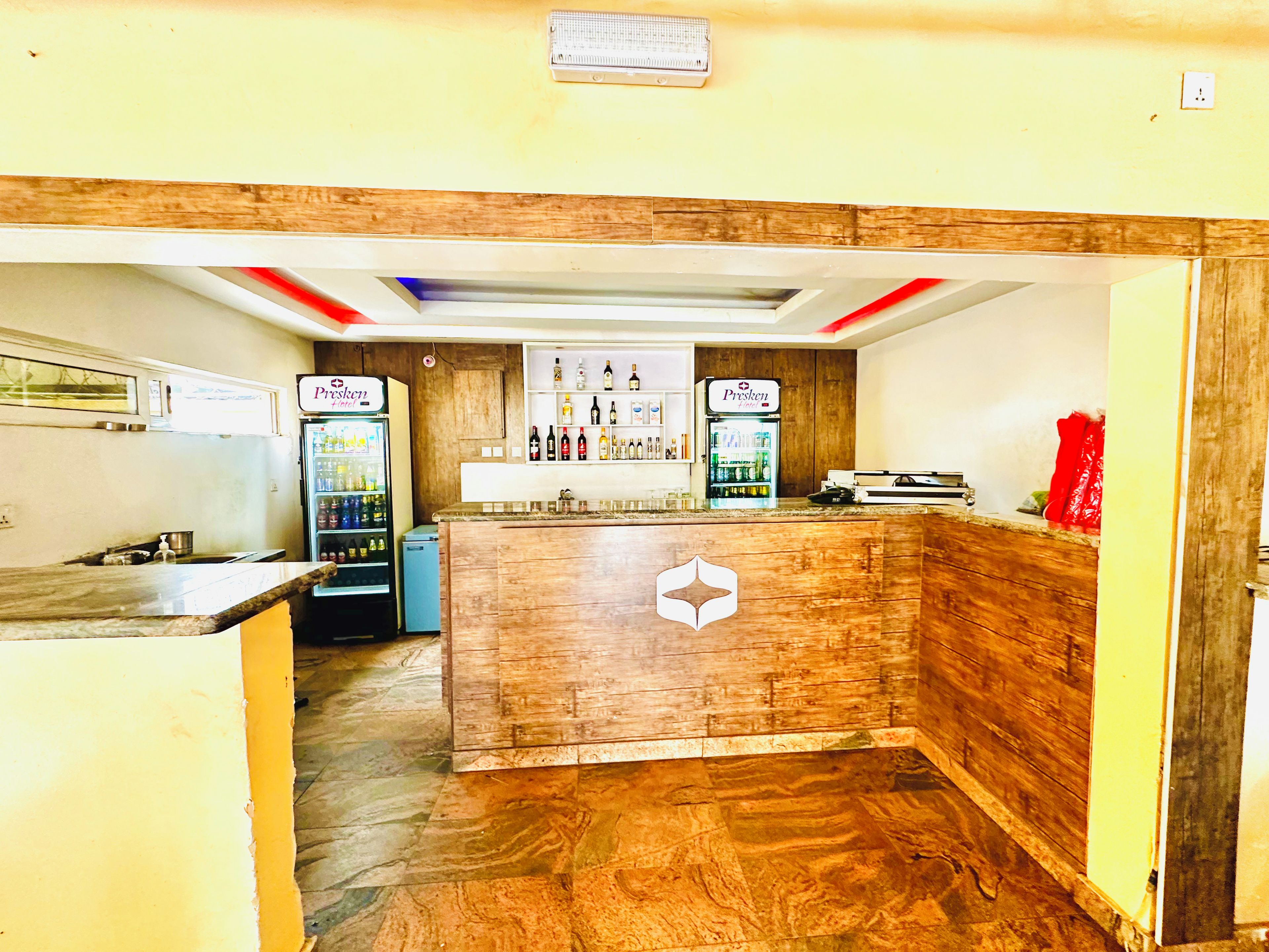 Un espace bar avec un comptoir en bois, deux réfrigérateurs remplis de boissons et une étagère fixée au mur où sont rangées des bouteilles.
