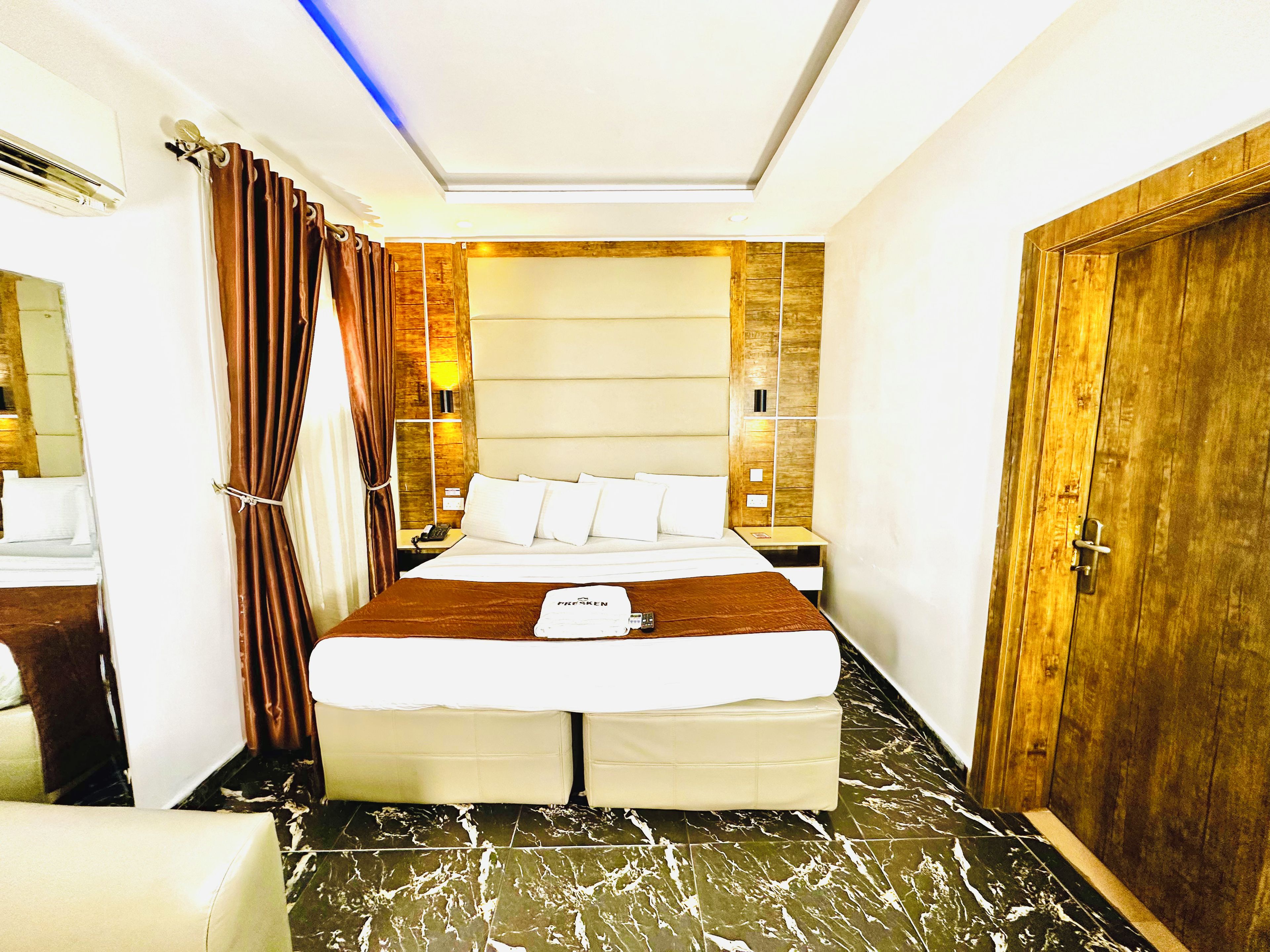 A hotel room with a large bed, a wooden wardrobe, a nightstand, and a bathroom visible in the background.