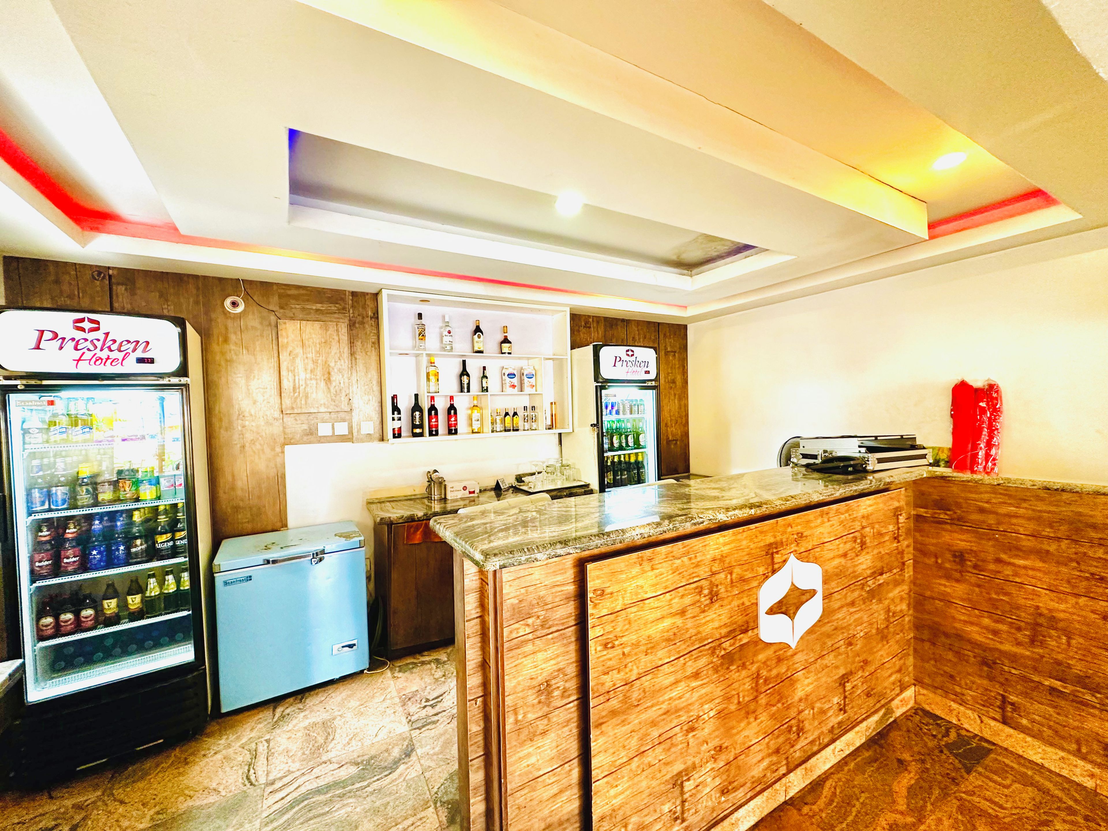 A hotel reception area with a wooden counter, two beverage dispensers, and a wall-mounted liquor display.