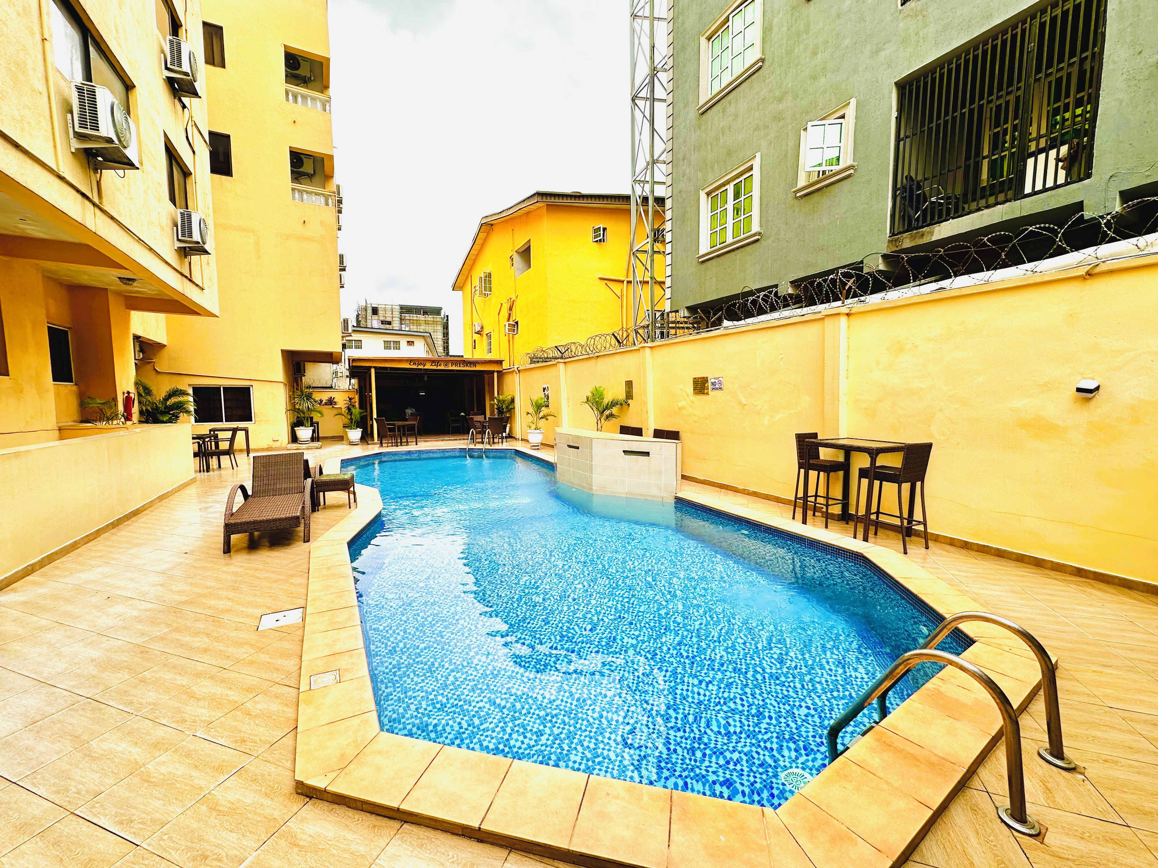 An outdoor swimming pool with a tiled deck, surrounded by yellow buildings, and furnished with outdoor seating.