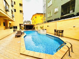 Piscina ao ar livre com deck ladrilhado, rodeada por edifícios amarelos e mobiliada com assentos externos.