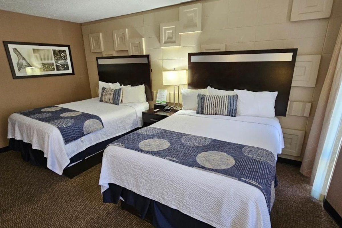 A hotel room with two beds, each with a headboard, a bedside table, and a lamp. The room has a carpeted floor and a framed picture on the wall.