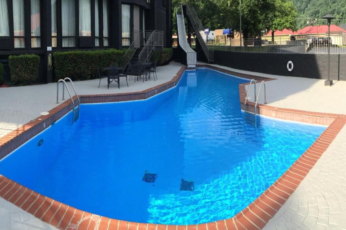 A swimming pool with a slide, surrounded by a brick border and a paved deck.