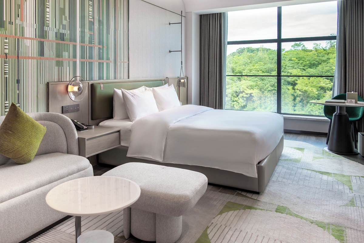 A modern hotel room with a bed, a sofa, a small table, and a view of the outdoors.