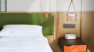 Una cama bien tendida con sábanas blancas, un cabecero verde, una mesita de noche con una lámpara y una maleta pequeña naranja y negra.