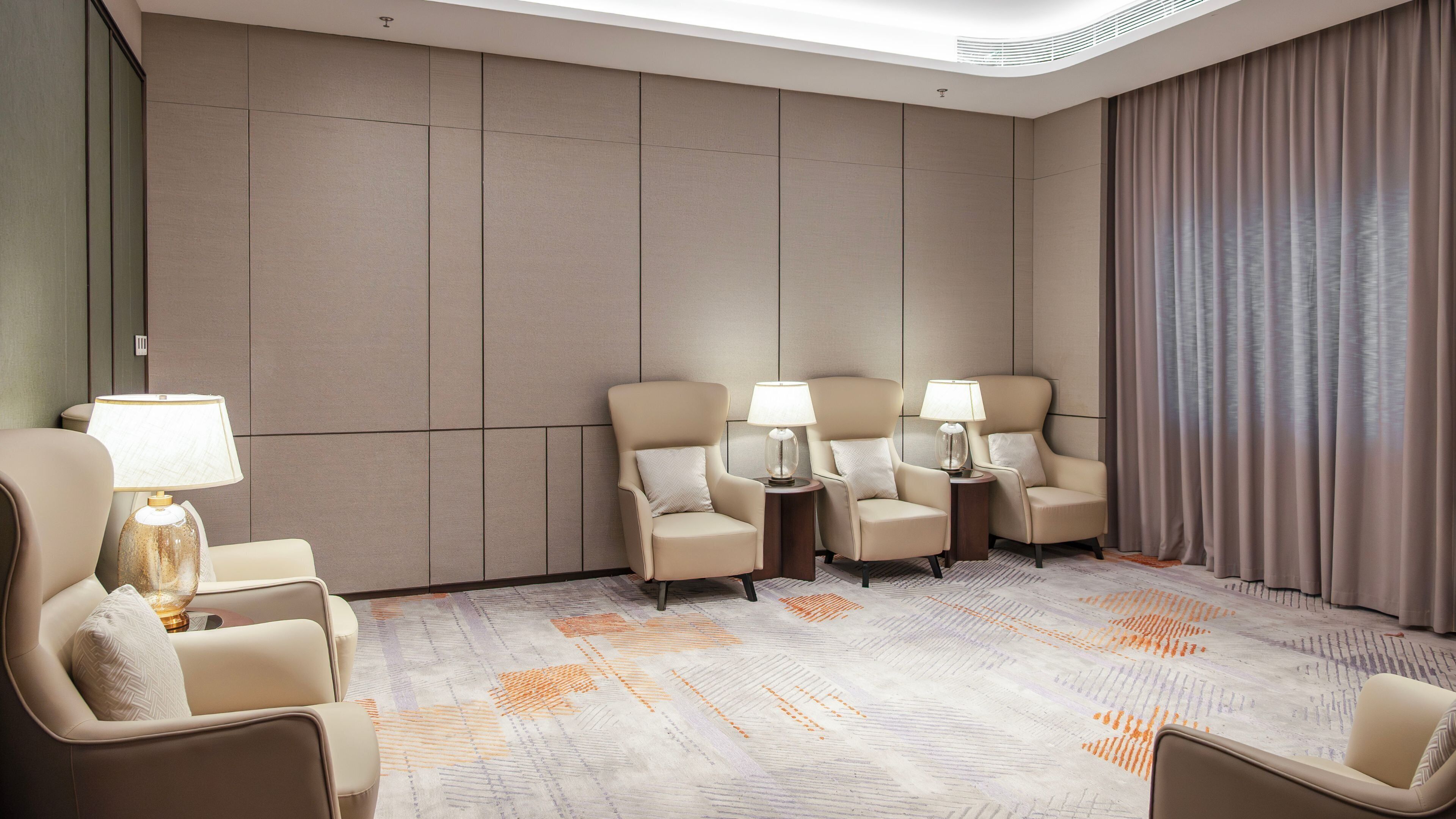Een hotelkamer met een groot raam, een bank, twee fauteuils, een kleine tafel met lamp en een kamerbreed tapijt met dessin.