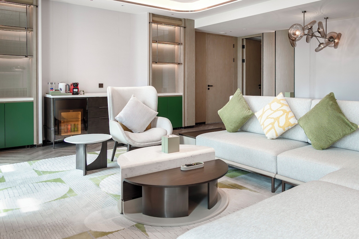 A modern living room with a sofa, armchair, coffee table, and a small dining area with a refrigerator.