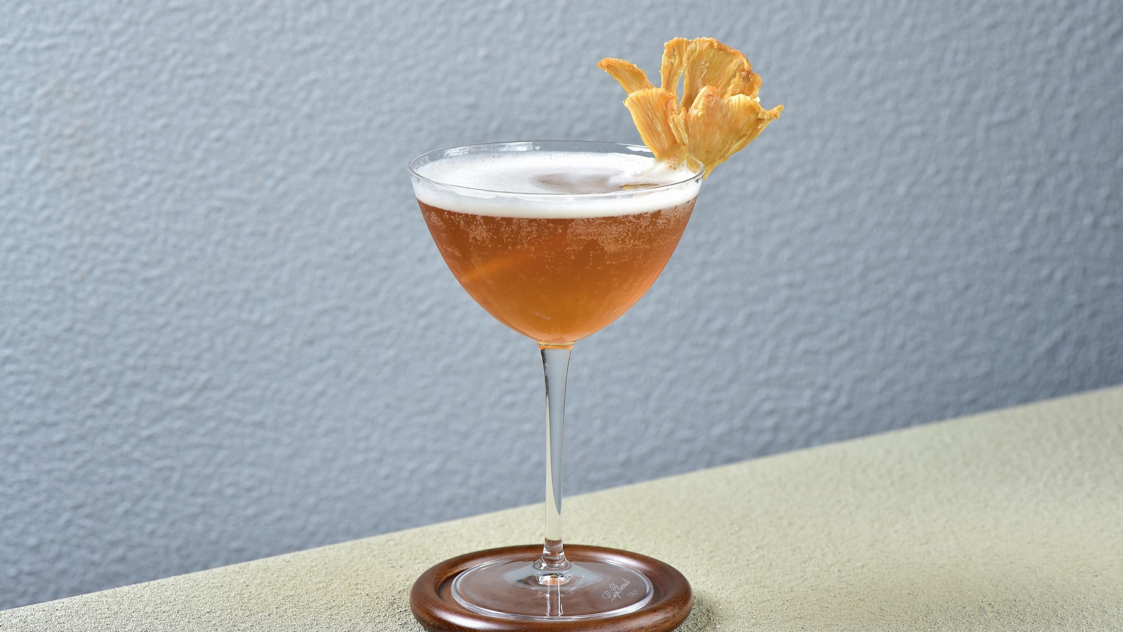 Un cocktail in un bicchiere da gambo con una guarnizione su un sottobicchiere di legno.
