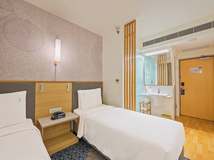 Habitación de hotel con dos camas individuales, una mesita de noche con lámpara, un espejo, una puerta de madera y un baño con lavabo y espejo.