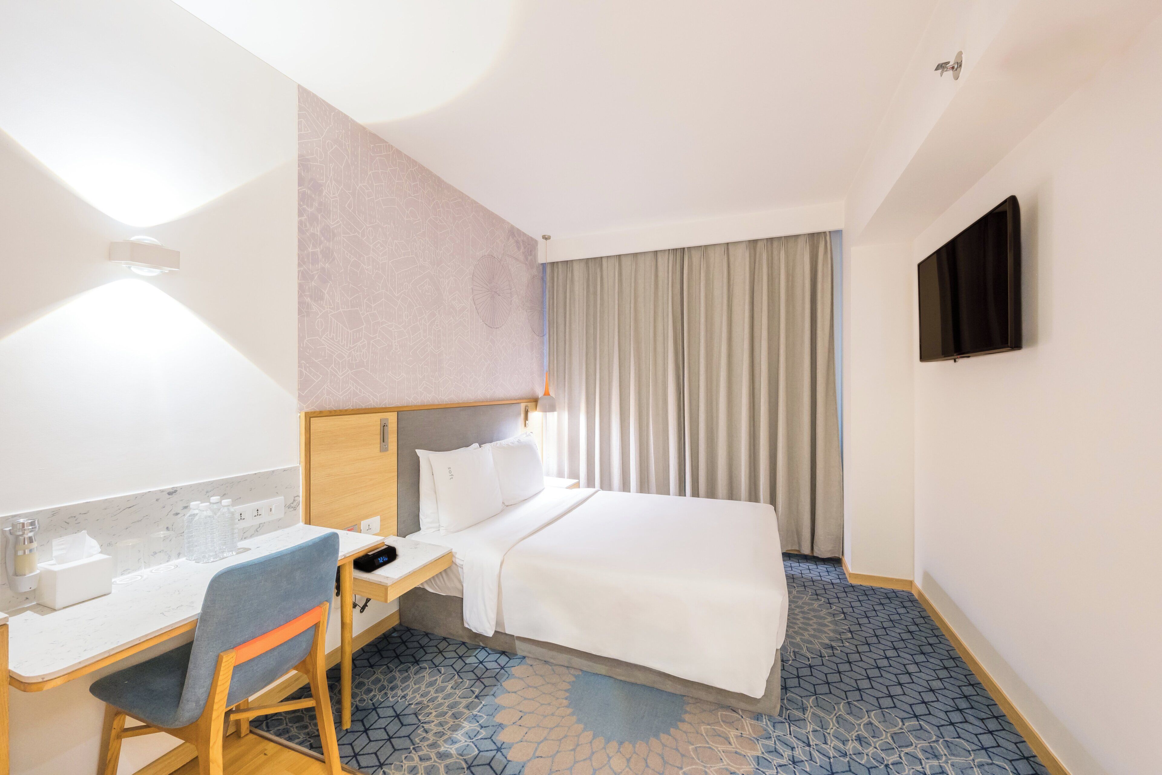 Una habitación de hotel con cama, escritorio con silla, televisor montado en la pared y ventana con cortinas.