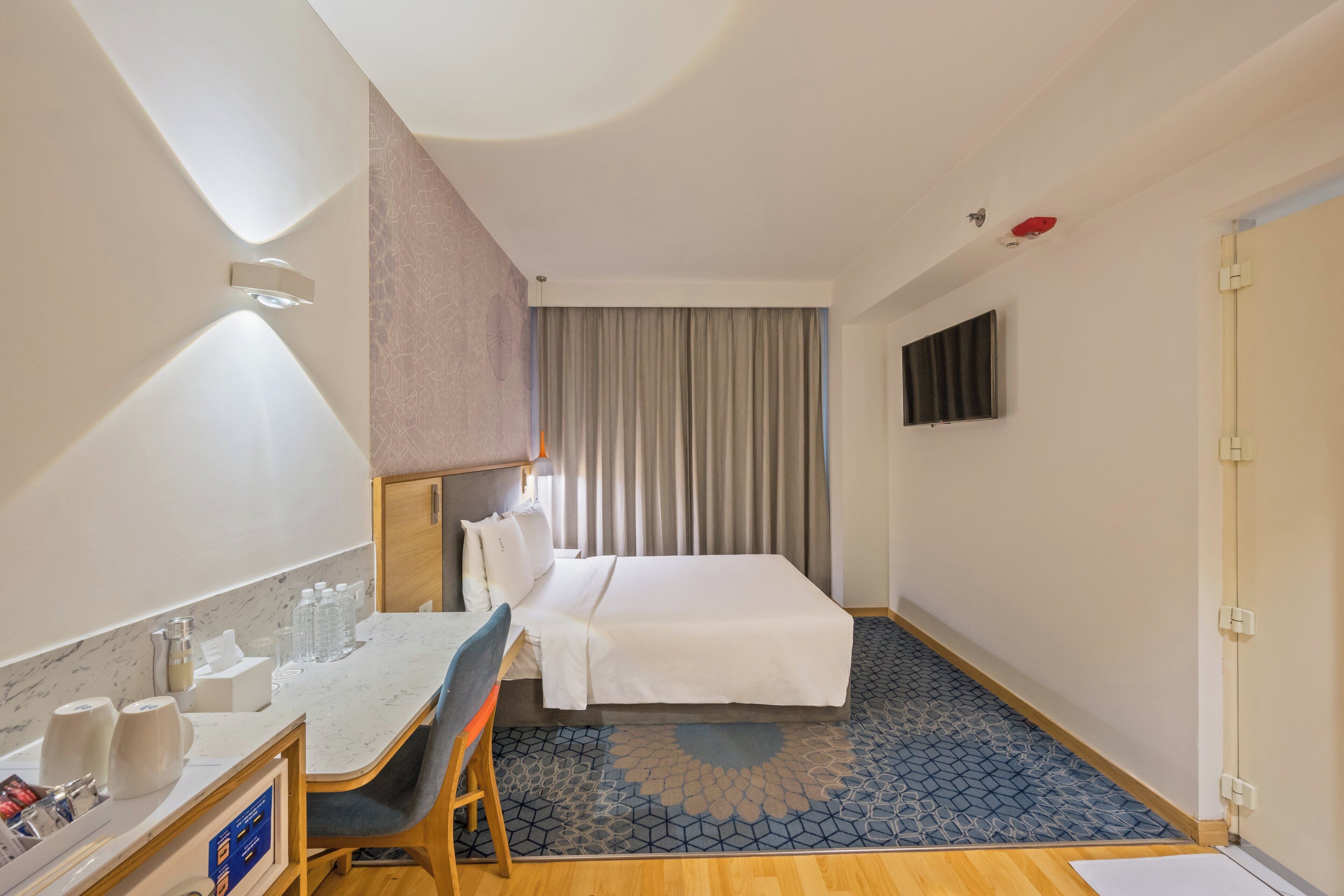A hotel room with a bed, a desk with a chair, a TV mounted on the wall, and a patterned rug.