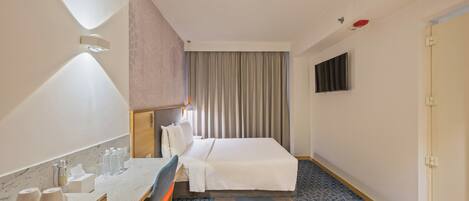 A hotel room with a bed, a desk with a chair, a TV mounted on the wall, and a patterned rug.