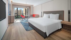 Une chambre d’hôtel moderne avec un grand lit, un balcon agrémenté d’une chaise rouge et une vue sur des immeubles de grande hauteur.