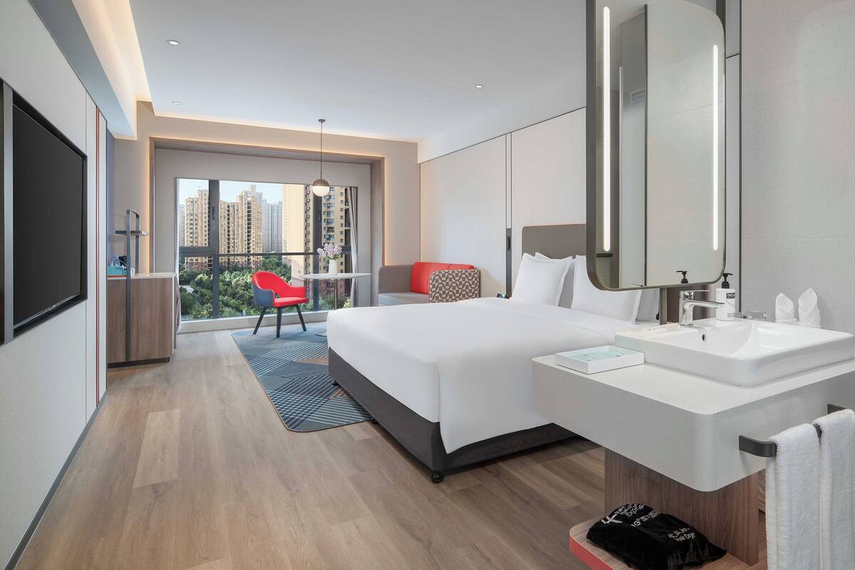 A modern hotel room with a large bed, a bathroom with a large mirror, and a view of the city through the window.