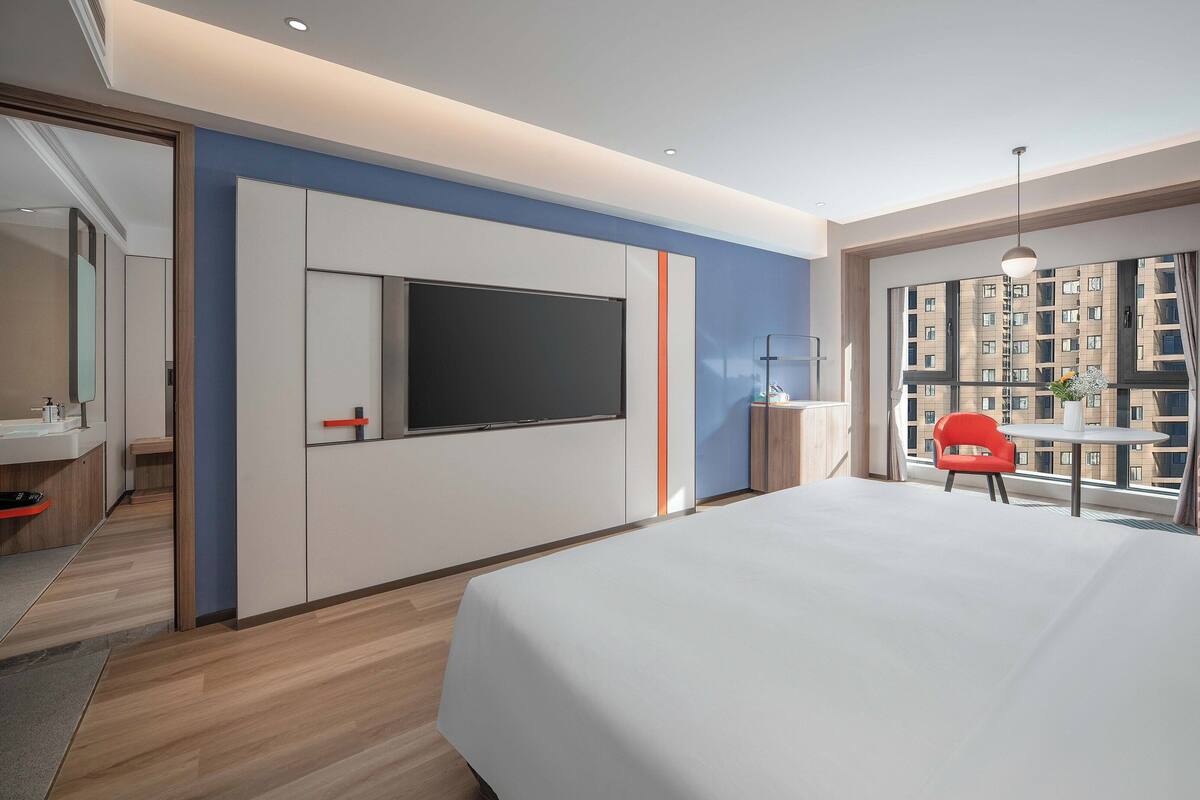 A modern hotel room with a large bed, a TV mounted on the wall, a small table with a chair, and a window with a city view.