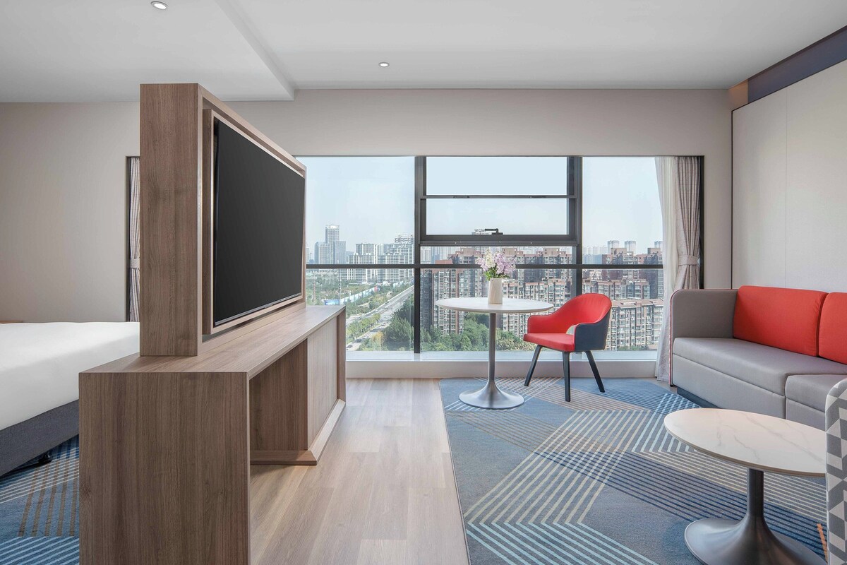 Een moderne hotelkamer met een groot bed, een televisie bevestigd aan een houten kastje, een rode fauteuil, een grijze bank en een balkon met uitzicht over de stad.