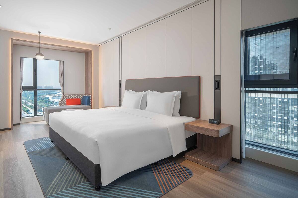 A modern hotel room with a large bed, a bedside table, a nightstand, a TV, and a view of the city through a large window.