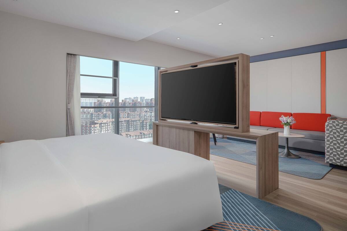 A modern hotel room with a large bed, a flat-screen TV, a sofa, and a view of the cityscape.