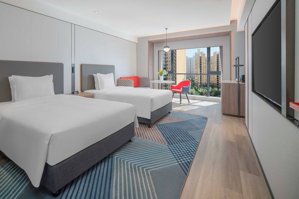 A modern hotel room with two beds, a balcony with a red chair, and a city view.