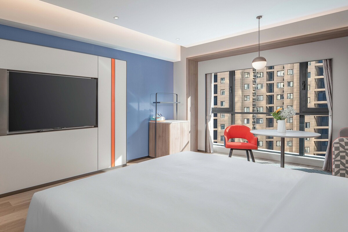 Habitación de hotel moderna con una cama grande, un televisor, una mesita con una silla y una ventana con vistas a la ciudad.