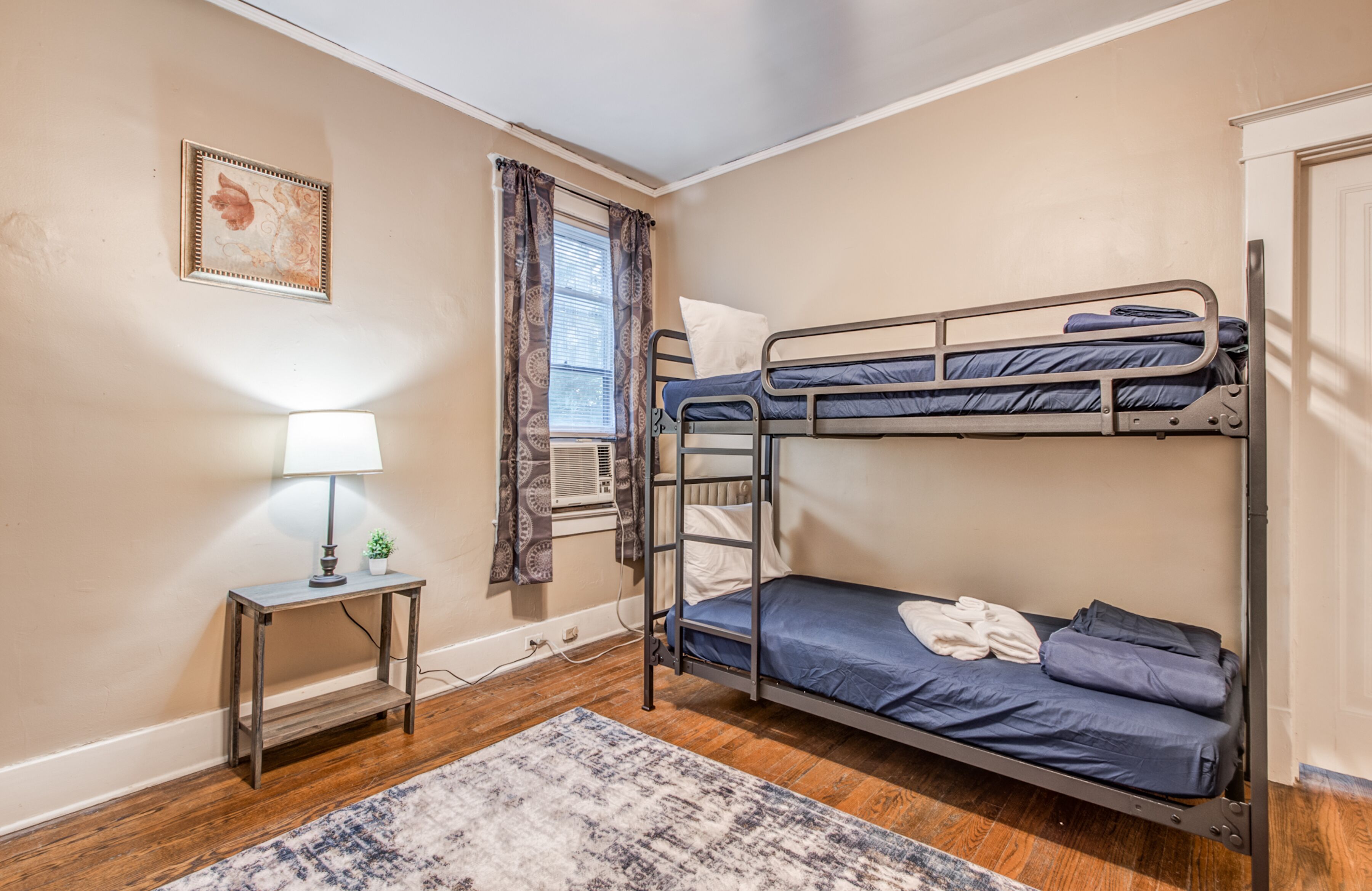 Quarto com beliche, mesa de cabeceira, luminária, janela com cortinas e tapete estampado.