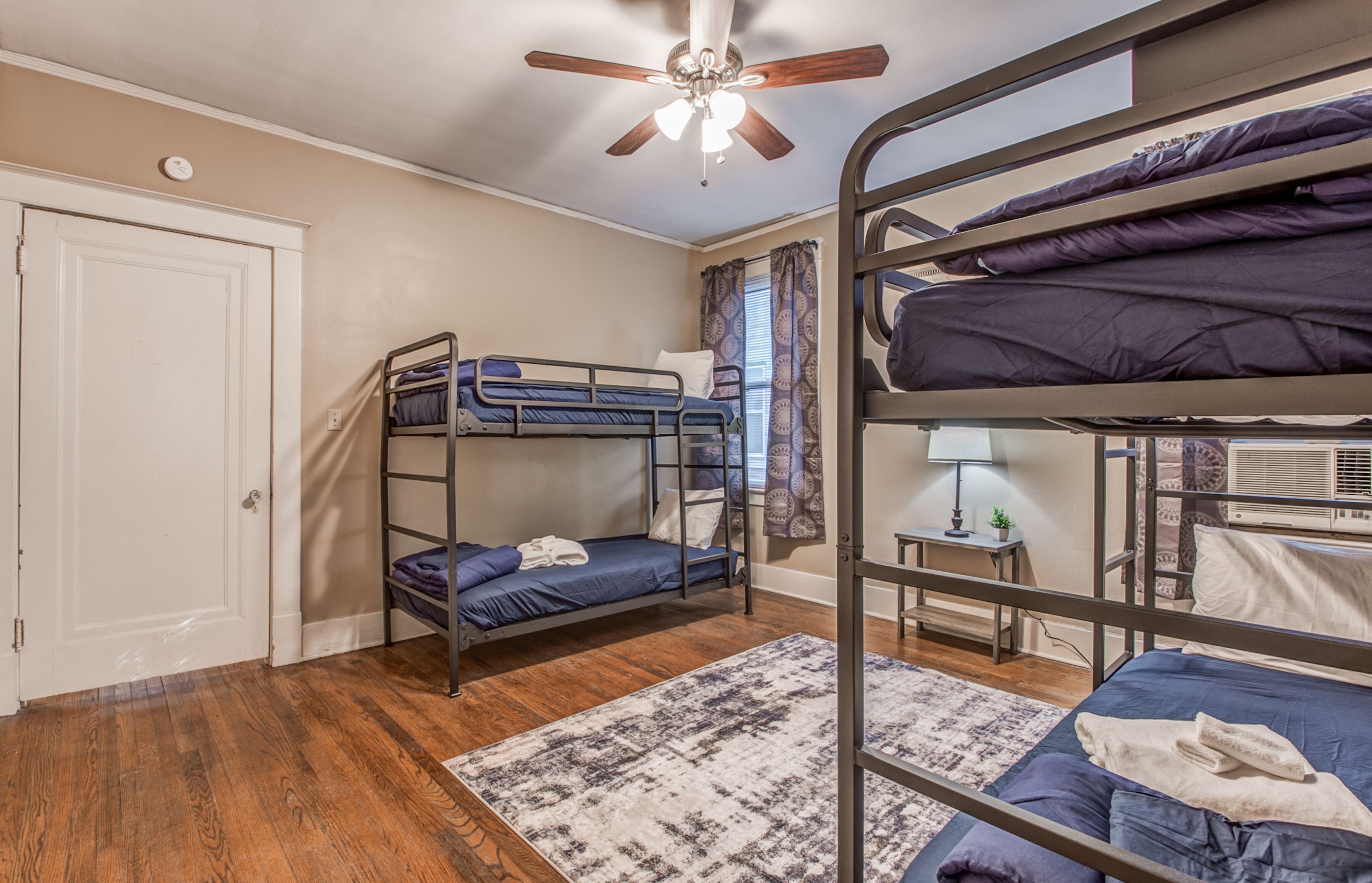 A dormitory room with bunk beds, a ceiling fan, a door, and a window with curtains.