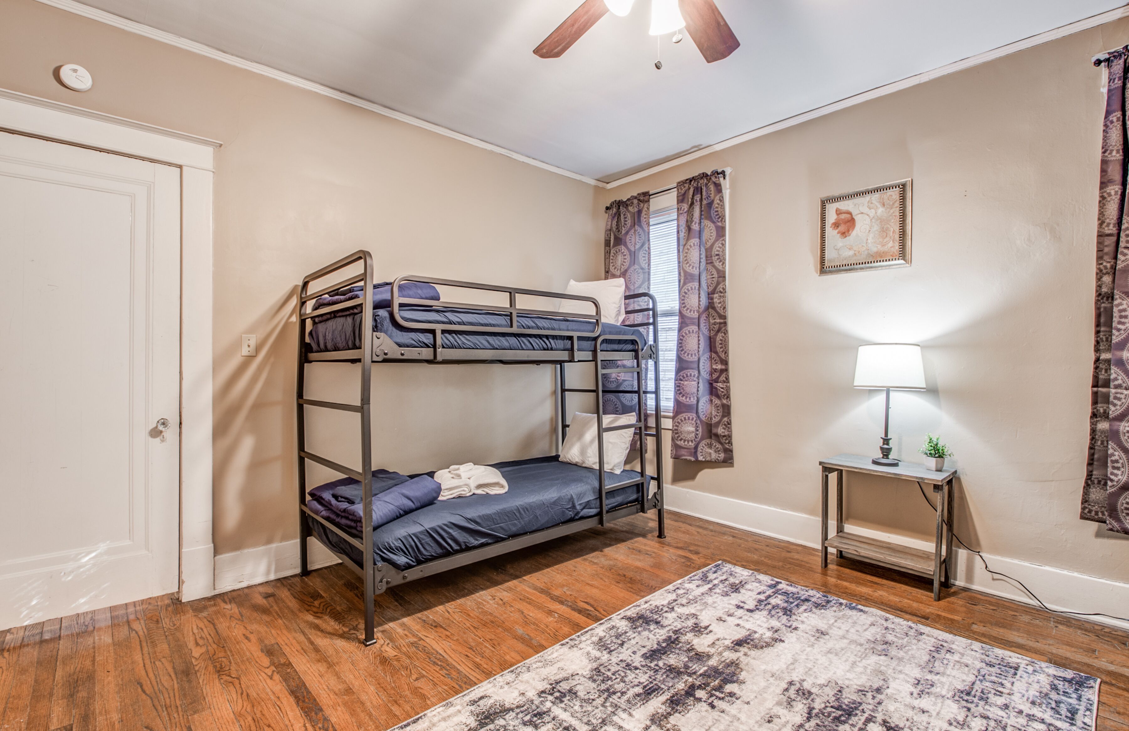 A bunk bed room with a wooden floor, a small table with a lamp, and a patterned rug.