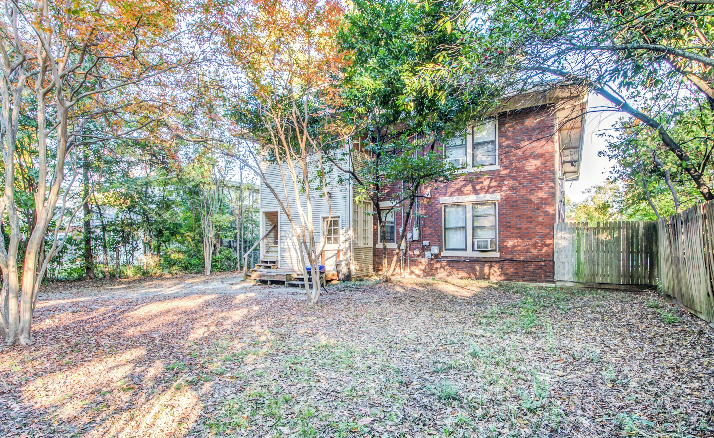 A residential backyard with a brick house, a wooden fence, and a gravel driveway.