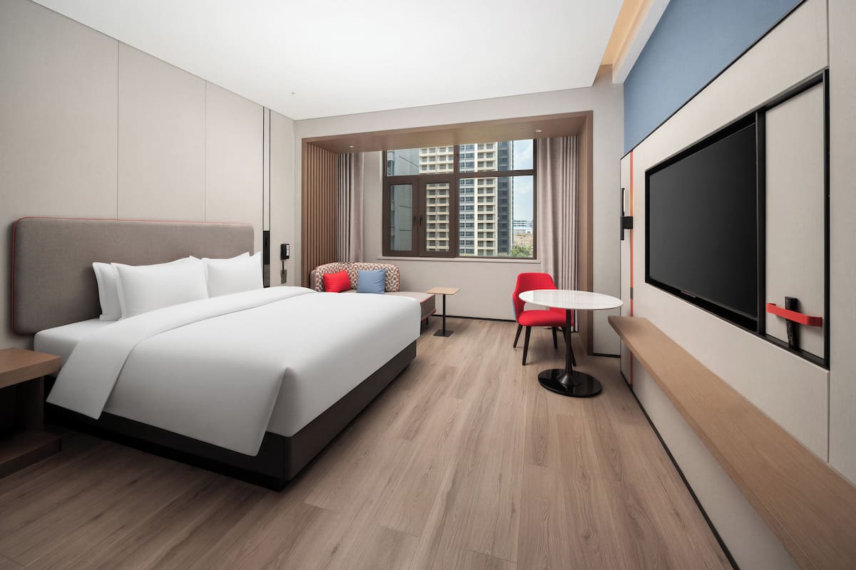 A modern hotel room with a large bed, a small table, a red chair, and a TV.