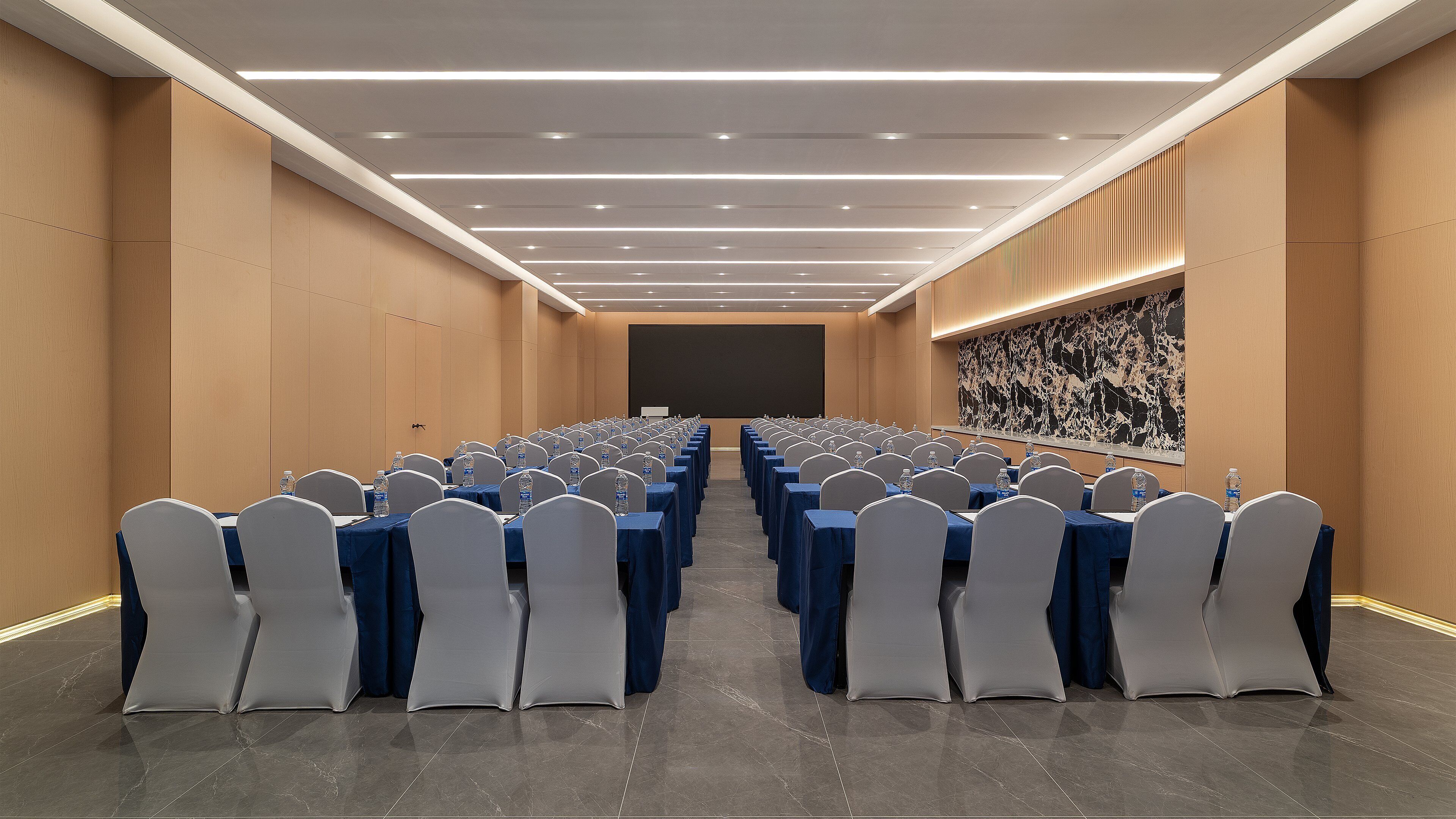 Una sala de conferencias con filas de mesas y sillas, una pantalla grande al frente y un dispensador de agua en cada mesa.