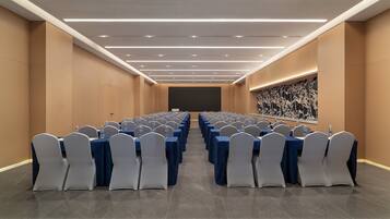 Una sala de conferencias con filas de mesas y sillas, una pantalla grande al frente y un dispensador de agua en cada mesa.