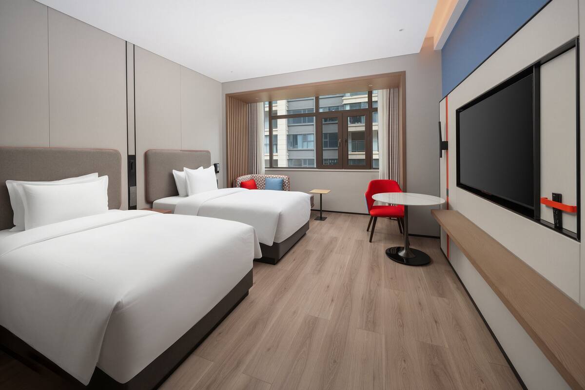 A hotel room with two beds, a TV, a small table, and a window with a view of buildings.