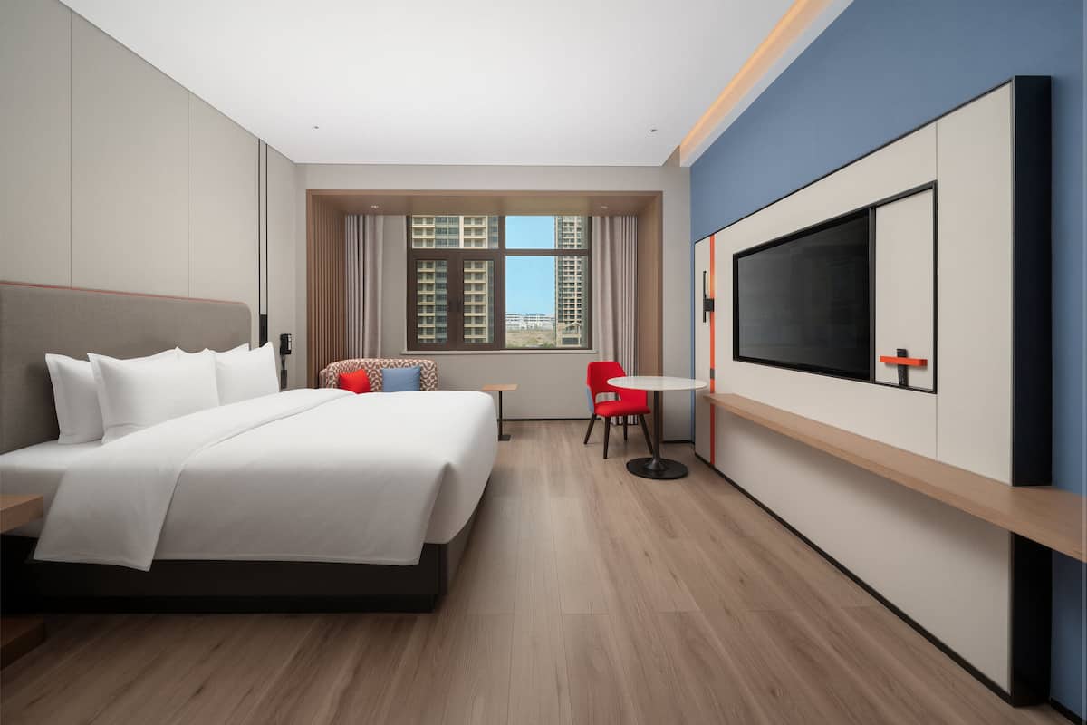 A modern hotel room with a large bed, a TV, a small table, and a window with a view of buildings.