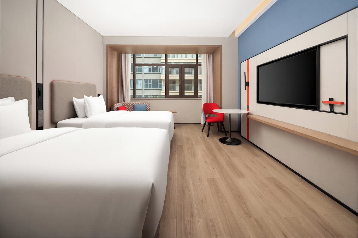 A hotel room with two beds, a TV, a small table, and a window with a view of buildings.