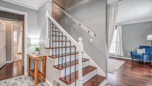 A staircase with wooden steps and black balusters, a blue armchair, and a white lamp on a wooden side table. - Shelby Parks Farm Haddington Home (Memphis)