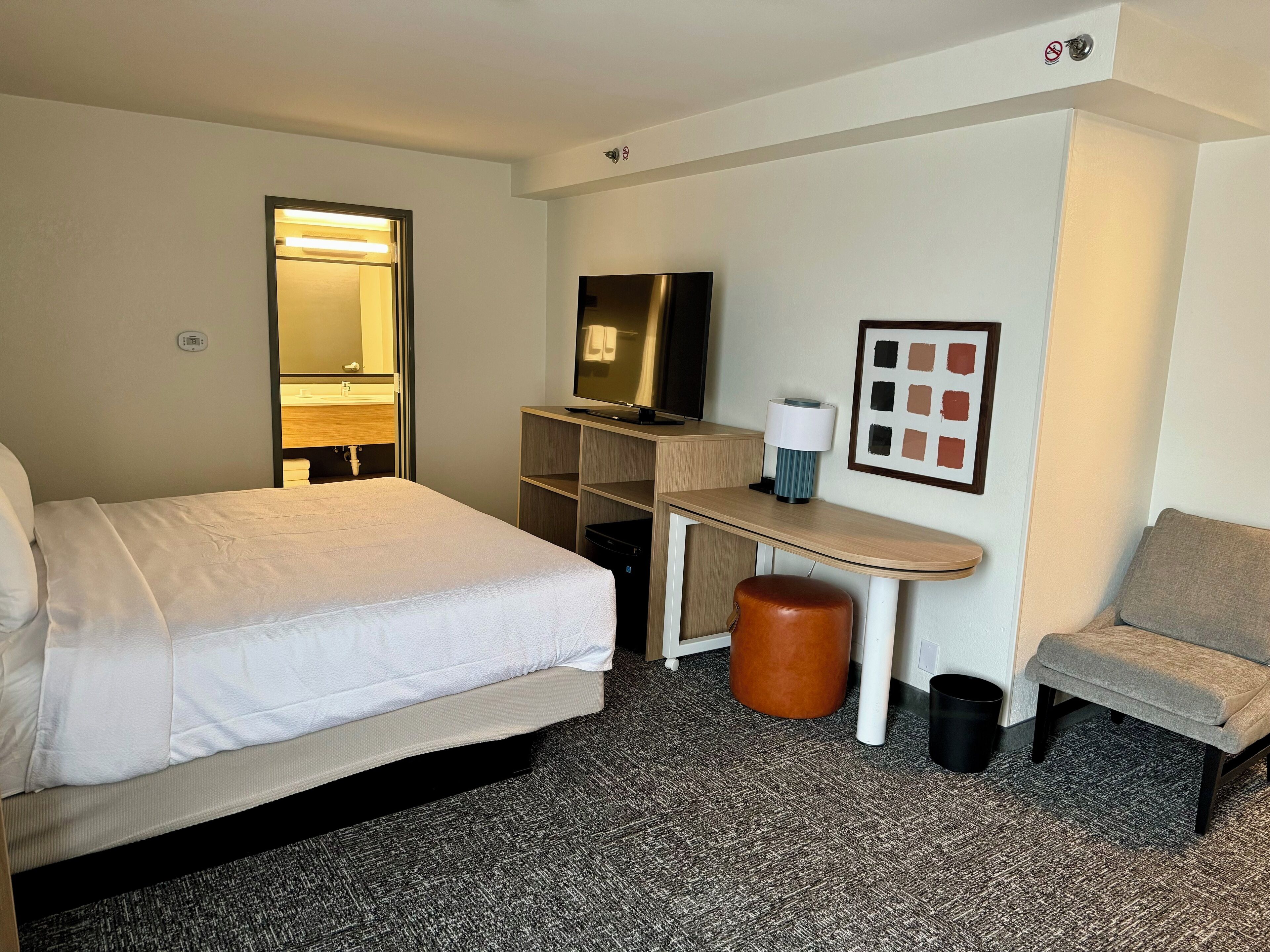 Une chambre d’hôtel avec un lit, un bureau avec un ordinateur, une chaise, une poubelle et un tableau encadré au mur.