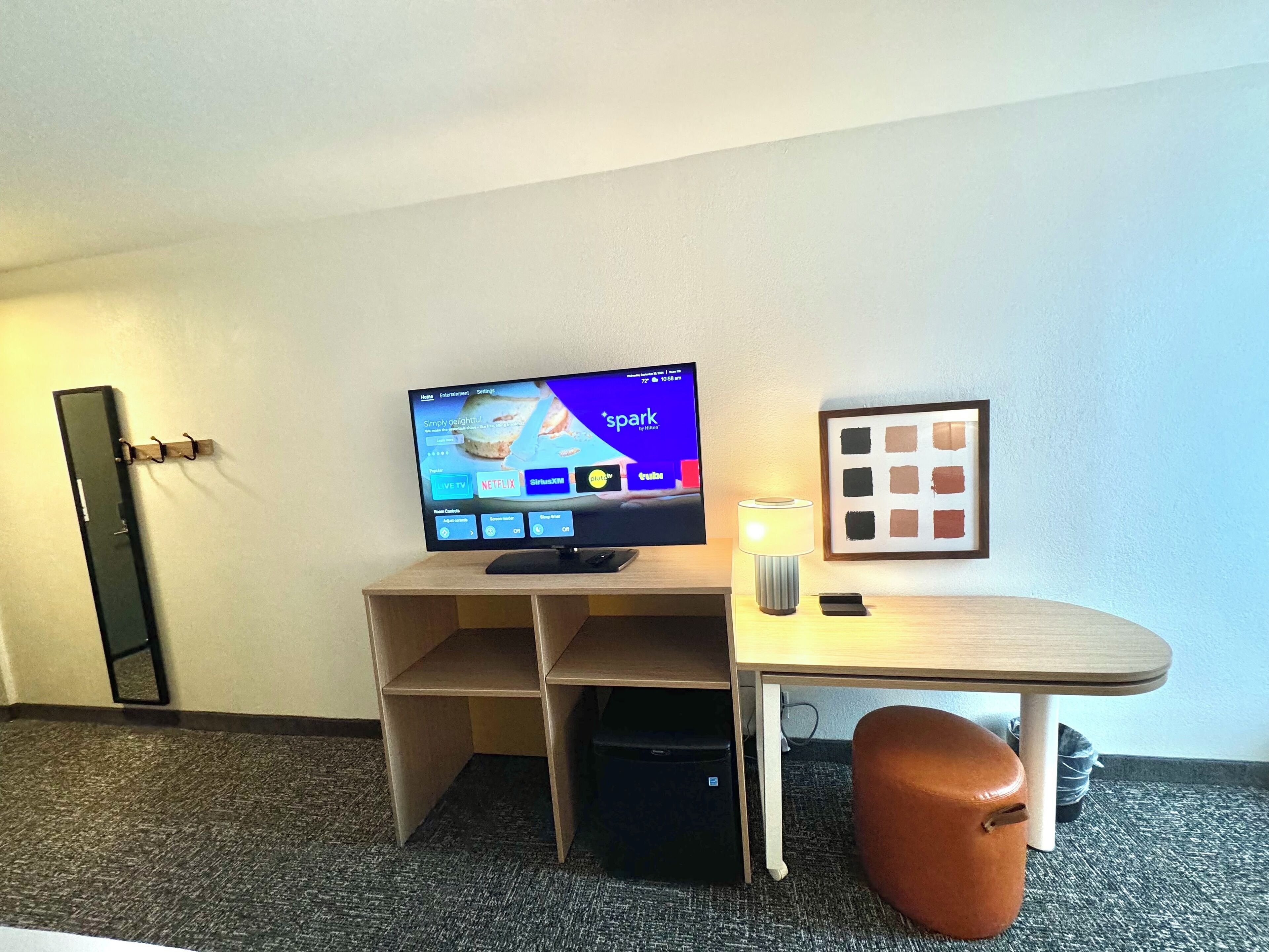 A hotel room with a TV, a desk, a lamp, a trash bin, and a framed picture on the wall.