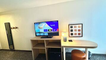 A hotel room with a TV, a desk, a lamp, a trash bin, and a framed picture on the wall.