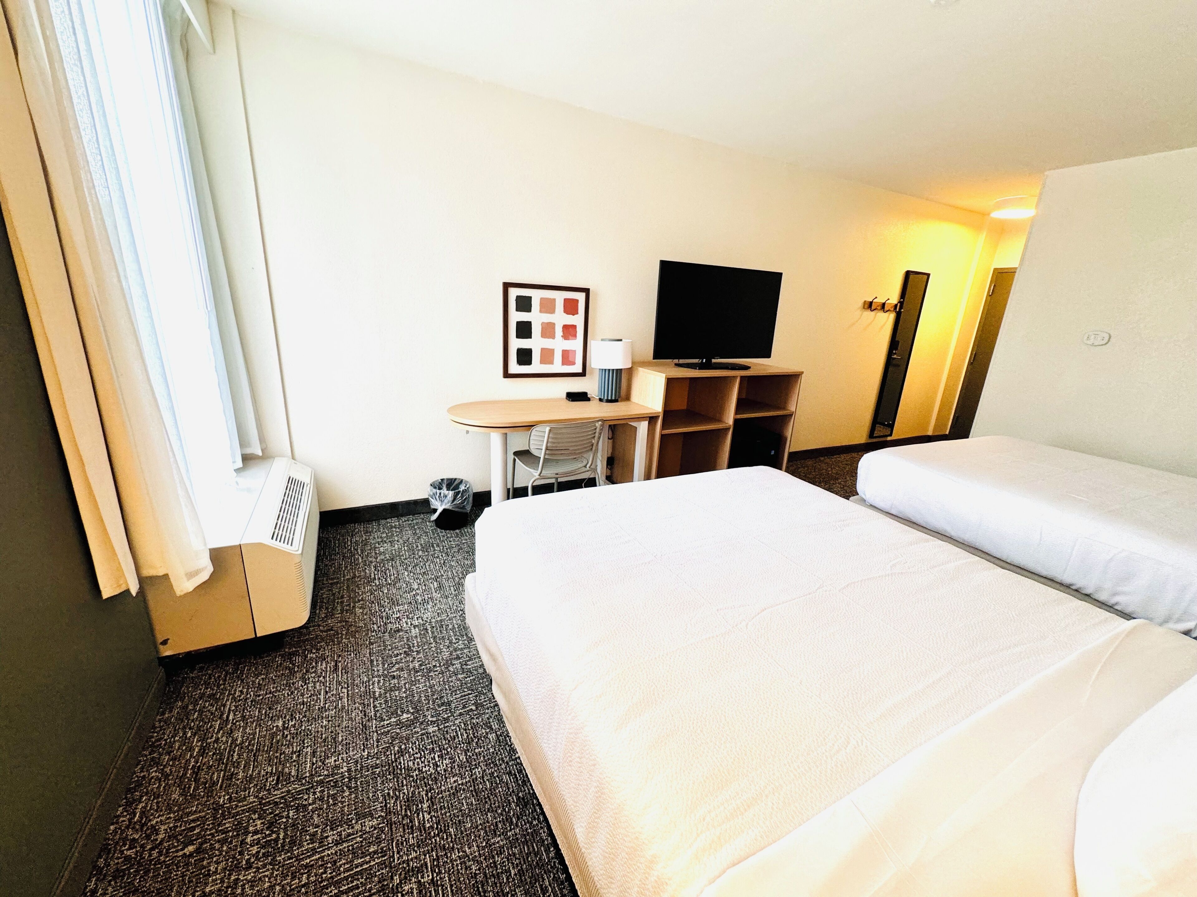 Habitación de hotel con dos camas, un escritorio con silla, un televisor y un cuadro en la pared.