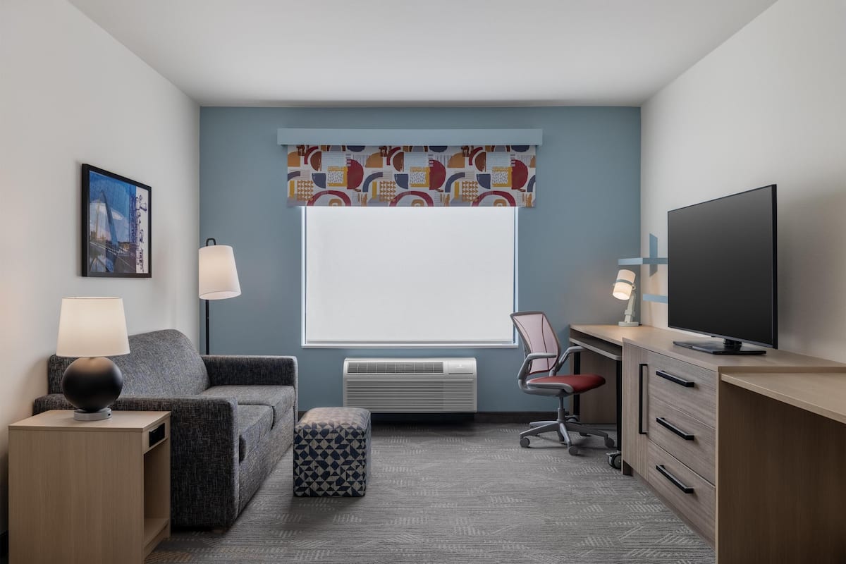 Een moderne hotelkamer met een grijze bank, een klein poufje, een bureau met een televisie en een raam met behang gordijnen.