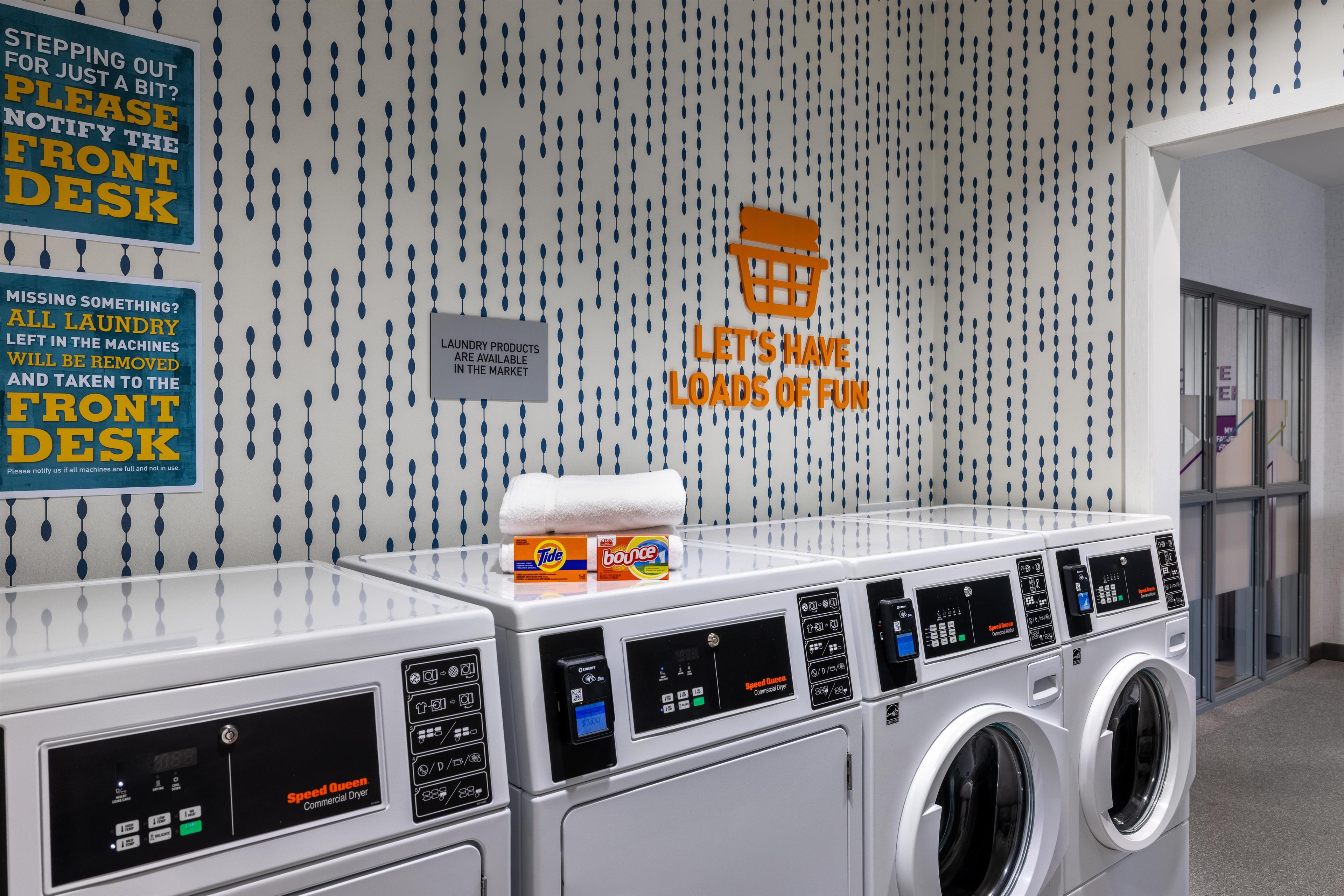 Zona de lavandería con varias lavadoras, un aviso sobre productos de limpieza y un cartel que invita a los clientes a disfrutar.