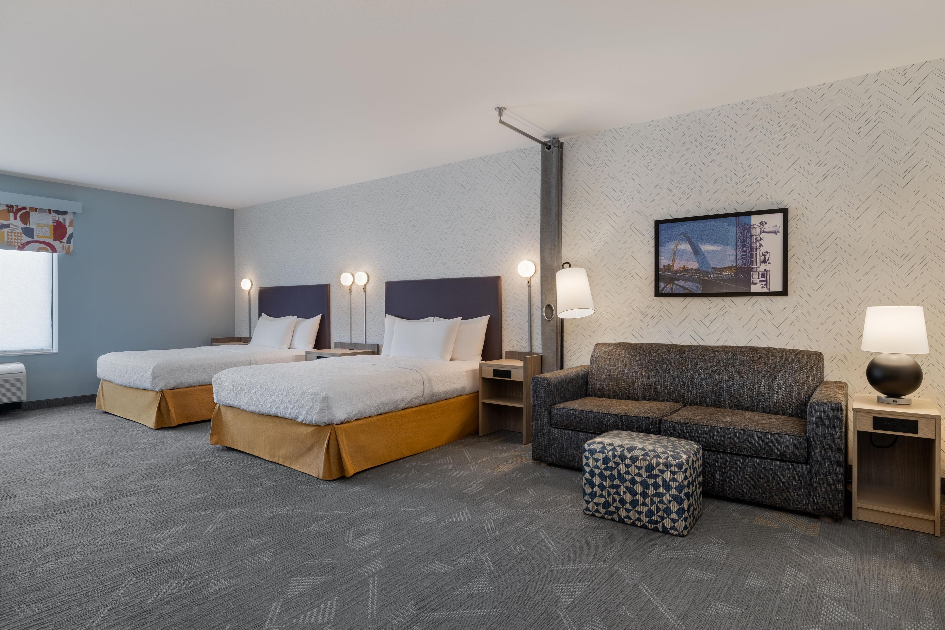 Une chambre d’hôtel avec deux lits, un canapé, un petit pouf et un tableau accroché au mur.