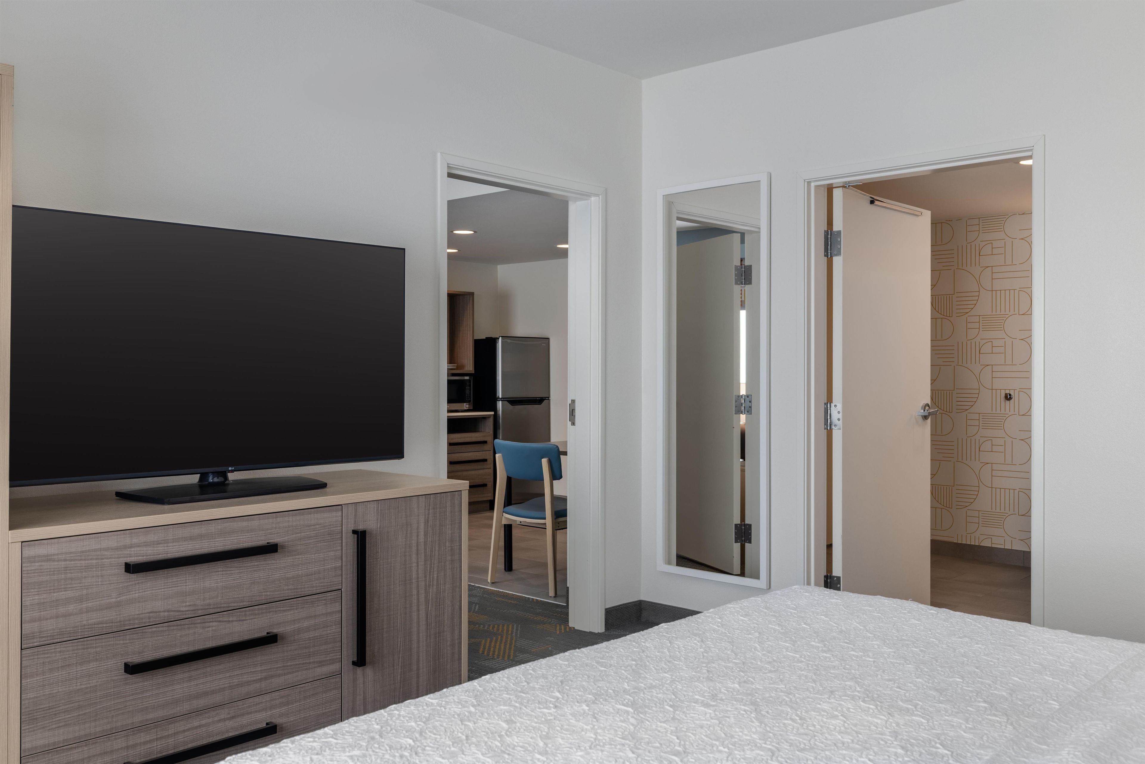 A hotel room with a flat-screen TV on a wooden dresser, a bed with a textured white cover, and a door leading to a bathroom with a patterned wall.