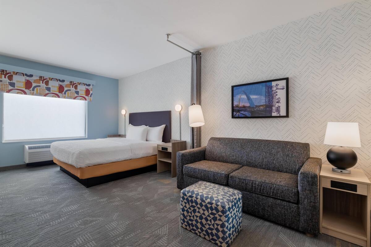 Hotelkamer met een bed, een bank, een gestreept voetenbankje, een nachtkastje met een lamp en een foto aan de muur.