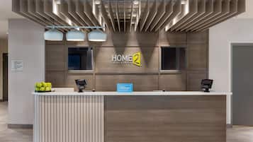 Una recepción de hotel con diseño moderno, que incluye un panel de pared de madera, un letrero de 'HOME 2 Suites by Hilton' y dos quioscos digitales.