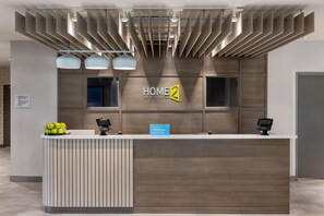 A hotel reception desk with a modern design, including a wooden panel wall, a sign for 'HOME 2 Suites by Hilton', and two digital kiosks. - Home2 Suites By Hilton Altoona Des Moines (Altoona)