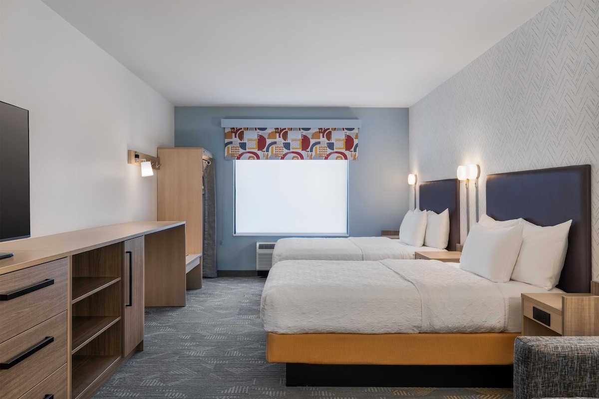 A hotel room with a bed, bedside tables, a desk, a wardrobe, and a large window with a view.